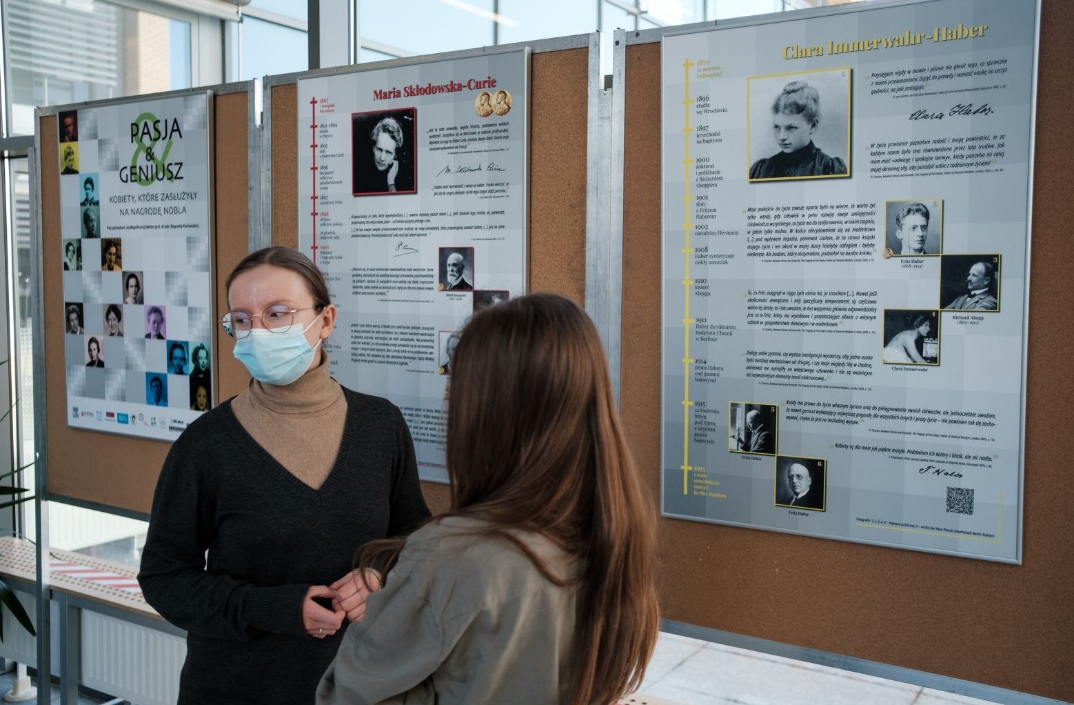 na zdjęciu dwie kobiety w maseczkach na tle kolejnych plakatów wystawy