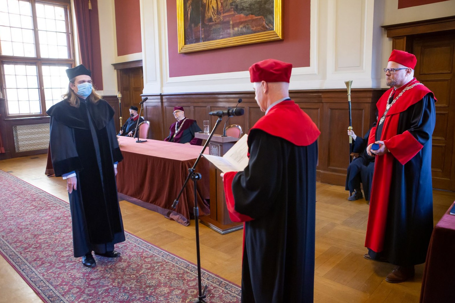 na środku sali stoi ostatnia z promowanych osób słuchając tekstu czytanego przez promotora , władze obok